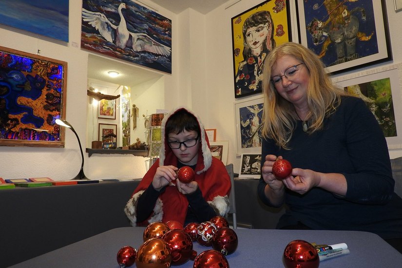 Weihnachtskind Hannes (li.) übt sich mit der Galerie-Gründerin Brigitta M. Arnold im Bemalen von Weihnachtskugeln vor den Gemälden von Frank-Ole Haake (li.) und Antje Menzel (re.).