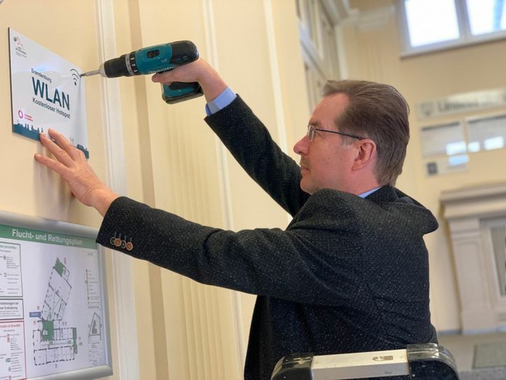 Das kostenfreie öffentliche Brandenburg-WLAN ist nunmehr auch an verschiedenen Standorten der Kreisverwaltung verfügbar. Bau-Dezernent Wilfried Brödno brachte das Hinweisschild im Landratsamt in Senftenberg im Bereich der Sitzungssäle an. Foto: Landkreis/Werner