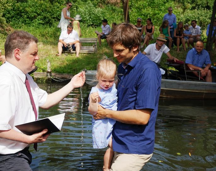 Die zweijährige Edda aus Hamburg fühlt sich bei ihrem Vater Christian sicher, als sie von Pfarrer Andreas Döhle am Samstag mit Spreewasser getauft wird. Für Mutter Sarah, die auf dem Grundstück an der Spree aufwuchs, ist die Spreetaufe ein Zeichen der Heimatliebe. Foto: Evangelischer Kirchenkreis Niederlausitz / Franziska Dorn