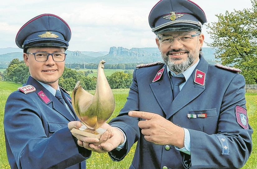 Die Feuerwehrmänner Toni Schulze (li.) und Kai Bigge mit der Goldenen Henne vor einem Panorama in der Sächsischen Schweiz.
