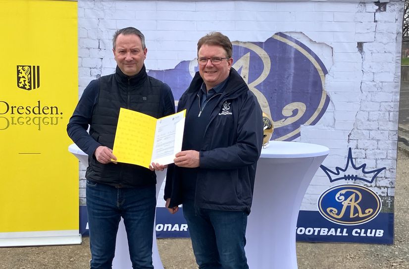 Sportbürgermeister Jan Donhauser (li.) übergibt vor dem Kunstrasenplatz den Fördermittelbescheid an den Präsident der Dresden Monarchs, Sören Glöckner.