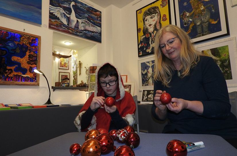 Weihnachtskind Hannes (li.) übt sich mit der Galerie-Gründerin Brigitta M. Arnold im Bemalen von Weihnachtskugeln vor den Gemälden von Frank-Ole Haake (li.) und Antje Menzel (re.).