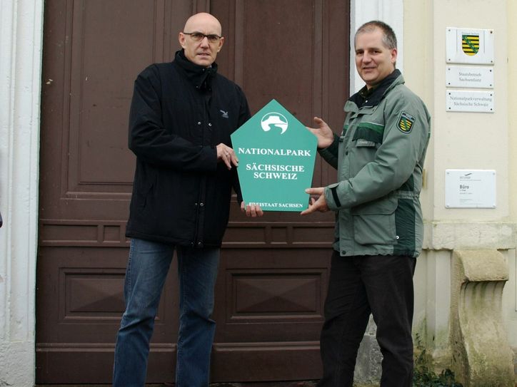 Mit der symbolischen Übergabe eines Nationalparkschildes überträgt Dr. Ingo Werners (l.) seine Aufgaben und Verantwortung für das Referat Betrieb/ Dienstleistungen an Michael Creutz (r.). Foto: Staatsbetrieb Sachsenforst