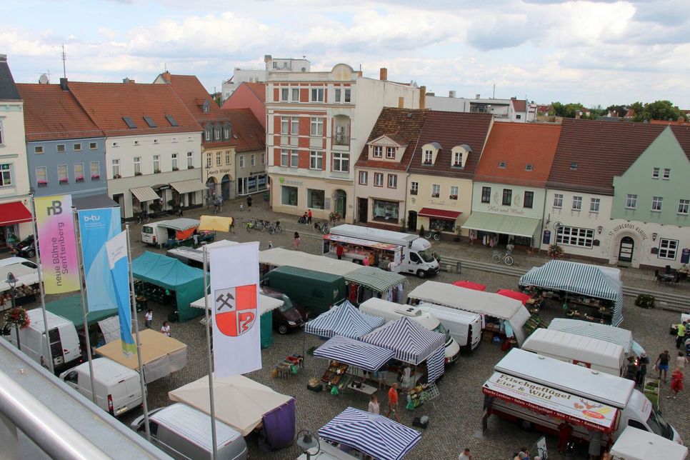 Blick auf den Marktplatz in Senftenberg. An der Westseite, hier direkt über der weißen Flagge mit dem Stadtwappen, könnte ein kleiner Baumhain aus sechs Zierkirschen in Baumkübeln entstehen.