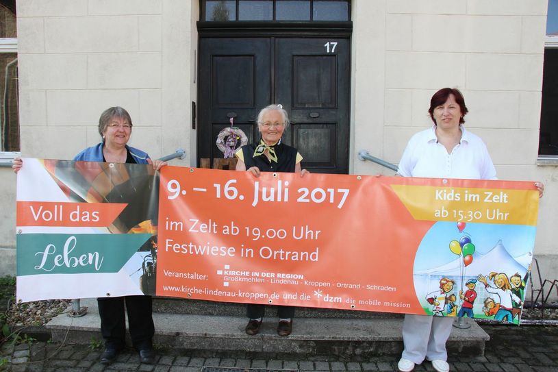 Freuen sich auf die Zelttage: Pfarrerin Angelika Scholte-Reh (li.), Gemeindesekretärin Monika Richter-Möllerke (re.) und Inge Merbeth (Mitte), eine von vielen Ehrenamtlichen im Dienst der Kirche. In der Hand halten sie ein Banner der Veranstaltung. Foto: S. Staindl