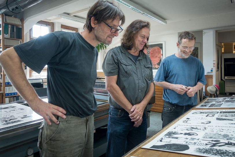 Drucker Torsten Leupold, Matthias Schroller, Künstler sowie Organisator des Projekts, und Werkstattleiter Peter Stephan, begutachten einen der ersten Abzüge der Postkarten-Edition, die anlässlich des 40. Dresdner Graphikmarktes in der Grafikwerkstatt Dresden entstanden sind.
